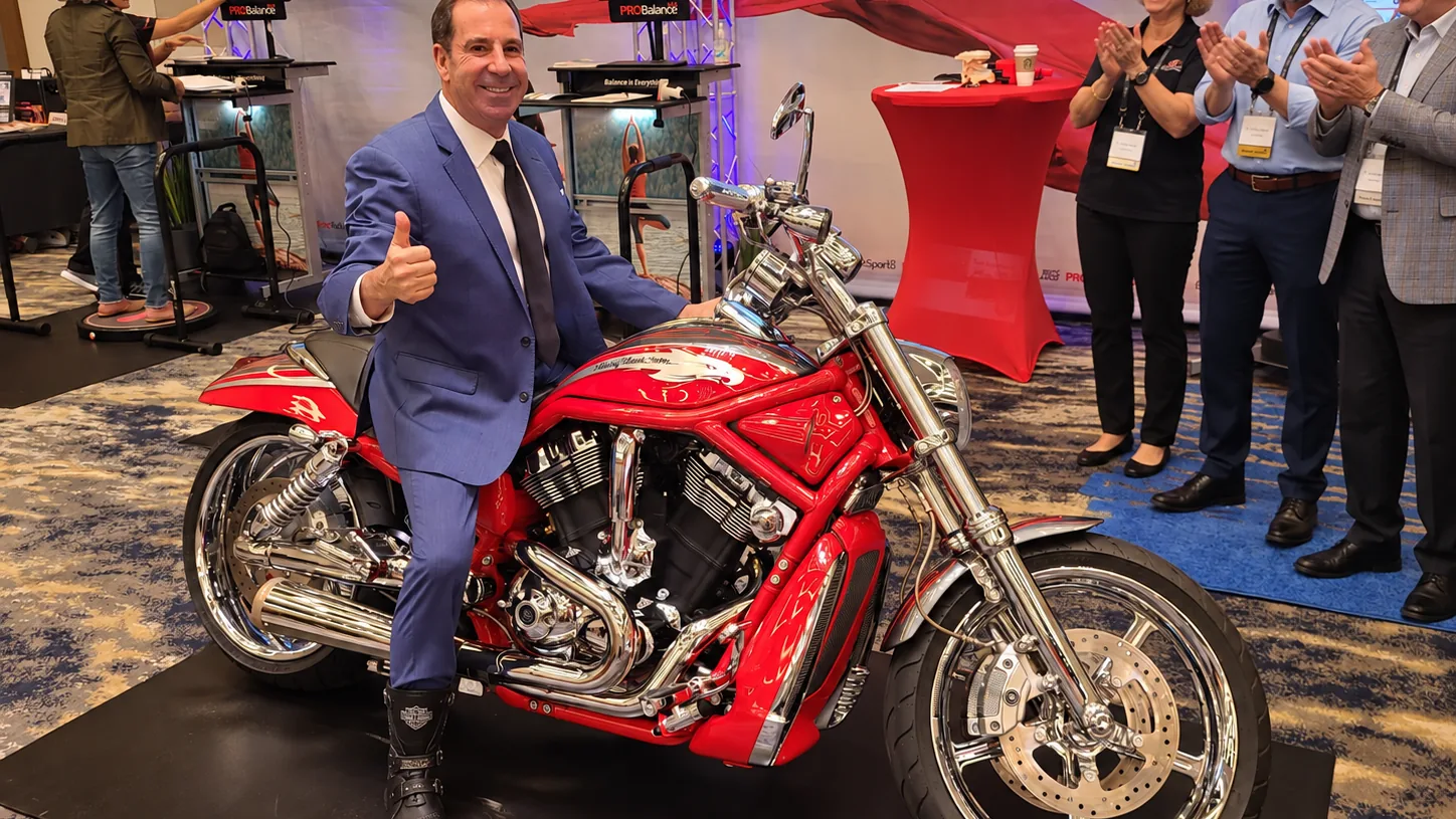 Dr. Mark Sanna giving a thumbs-up on a red motorcycle at the PRObalance exhibit booth
